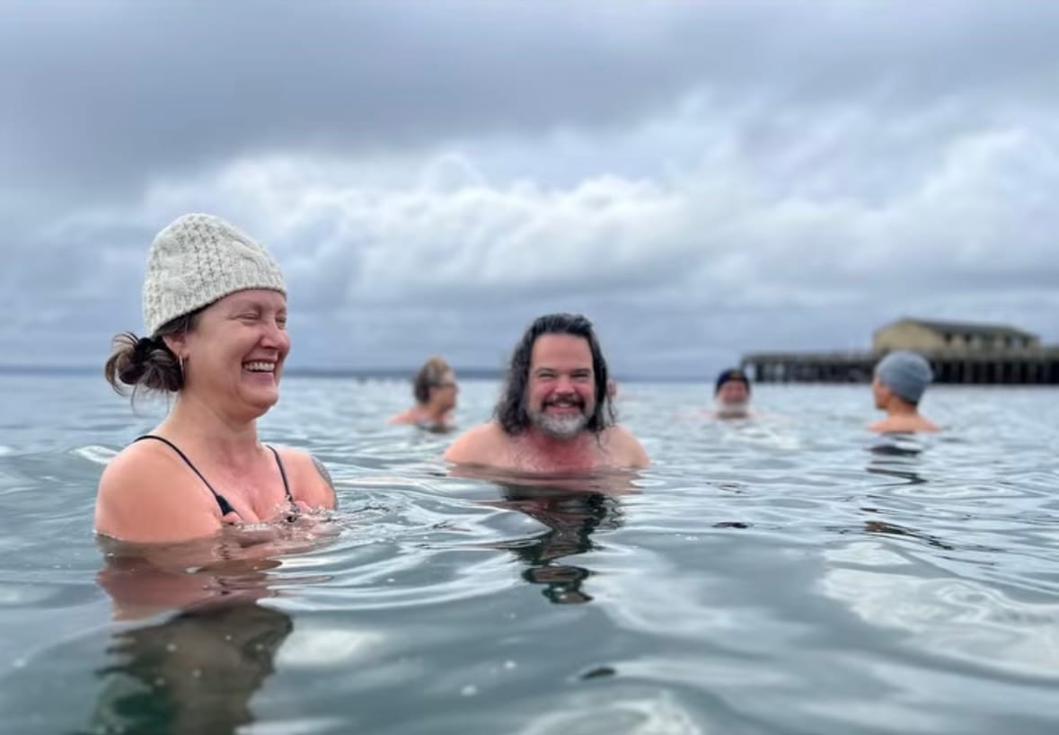 Five people up to their shoulders in the sea on a day with heavy clouds. Three of the people are wearing warm winter hats. Everyone is smiling.